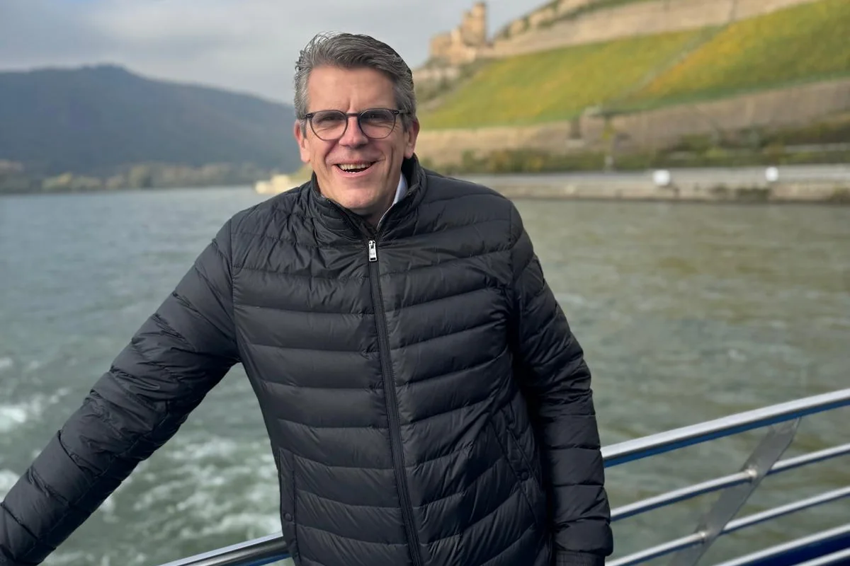 Günther Wagner auf einem Rheinschiff bei Rüdesheim, im Hintergrund Weinberge und eine Burg.